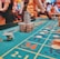 A casino scene with a roulette table features prominently, covered in green felt with colorful numbers and betting squares. Several stacks of poker chips are placed on different numbers, and the hands of several people are visible as they interact with the table. In the background, there are slot machines and more people, creating a vibrant and busy atmosphere.