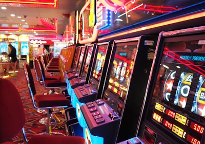 A brightly lit casino interior featuring a row of colorful slot machines. The neon lights create a vibrant atmosphere, with patterned carpeting adding to the decor. Stools are aligned neatly in front of each machine.