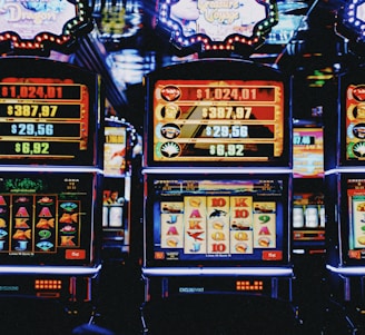 A row of colorful slot machines lit with bright neon lights, showcasing various graphical elements on the screens, indicating different games and jackpots. The atmosphere suggests a lively casino setting, bustling with excitement.