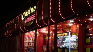A brightly lit casino lounge entrance adorned with neon lights. The exterior features colorful signage including the words 'Casino' and 'Lounge' in vivid colors, with additional lights framing the building. Inside, a glimpse of a claw machine filled with plush toys and arcade games is visible. The area exudes an inviting yet bustling atmosphere typical of entertainment venues.