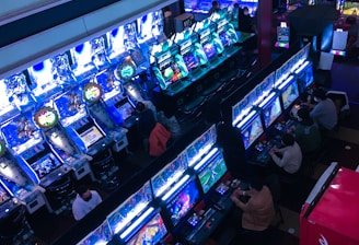 A bustling arcade filled with rows of gaming machines, each with vibrant displays and controls. Several people are seated at these machines, deeply engaged in playing games. Bright fluorescent lights cast a blue and neon glow across the room, enhancing the lively and energetic atmosphere.