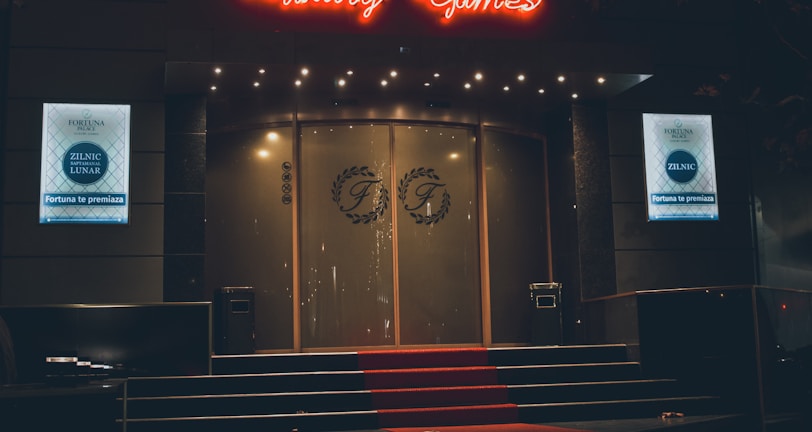 A neon-lit casino entrance with a red carpet leading up to large double doors. The sign reads 'Fortuna Palace Luxury Games' in bright red and white neon lights. The dimly lit atmosphere suggests a night-time setting. There are two digital displays on either side of the entrance showing promotional content.
